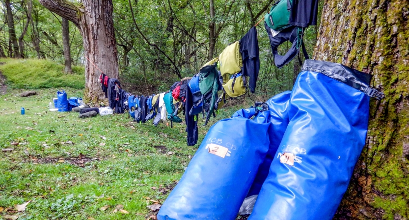 A clothesline tied between two trees holds gear that is drying. There are two dry bags resting against the tree in the foreground. 